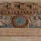 La complessit� della decorazione si infittisce nel fregio, dove foglie intrecciate conducono a uno stemma centrale azzurro acquerellato con la scritta Charitas
