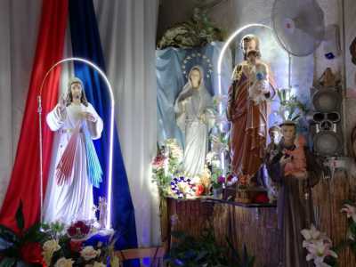 Nino ci accoglie subito sorridente e ci fa entrare nella stanza. Mentre veniamo accolti dal sottofondo di una musica religiosa notiamo due strette file di banchi poste davanti a un transetto di legno, oltre il quale svettano decine di grandi statue di santi, adornate di fiori profumati
