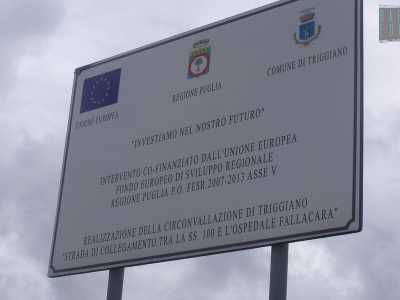 Un cartello recita trionfalmente: "Investiamo sul futuro". E dopo aver chiarito che si tratta di fondi europei per la Regione Puglia (2007-2013), ci segnala che quella abbiamo davanti � proprio la "Circonvallazione di Triggiano", precisamente la "strada di collegamento tra la SS 100 e l'ospedale Fallacara"
